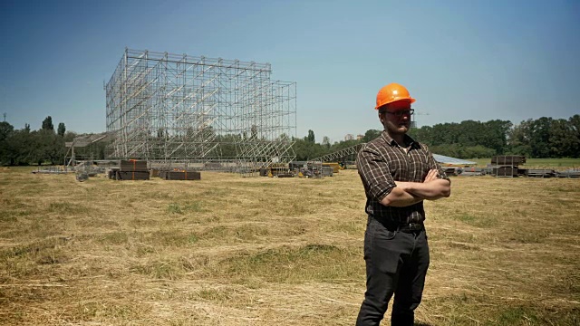 信心十足的建筑工人双手交叉站在头盔上，背景是稻草地的金属建筑视频素材