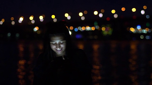 一个女孩开心地用智能手机站在一座夜城的背景上视频素材