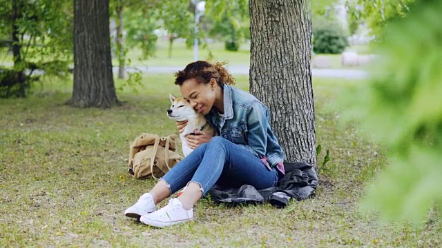 活泼的混血女孩拥抱着美丽的日本柴犬，抚摸着在公园树下休息的动物，而宠物享受着爱和照顾。视频素材