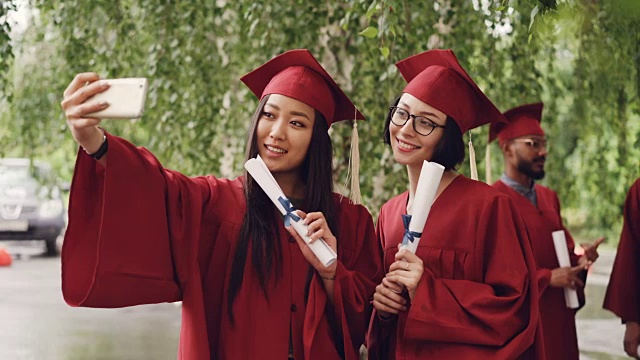 漂亮的女生们用智能手机和毕业证自拍，女生们在摆姿势、微笑，同学们在背景中庆祝。视频素材