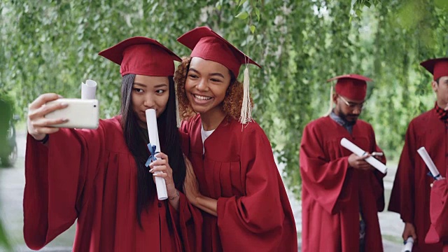 两个漂亮的女同学在毕业典礼上拿着毕业证书自拍，年轻的女同学在微笑、摆姿势、做手势、做鬼脸。视频素材