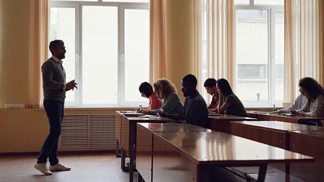 大胡子大学老师正在给成年学生打手势朗读讲座，年轻人则在写作和听他讲课。高等教育与青年观念。视频素材