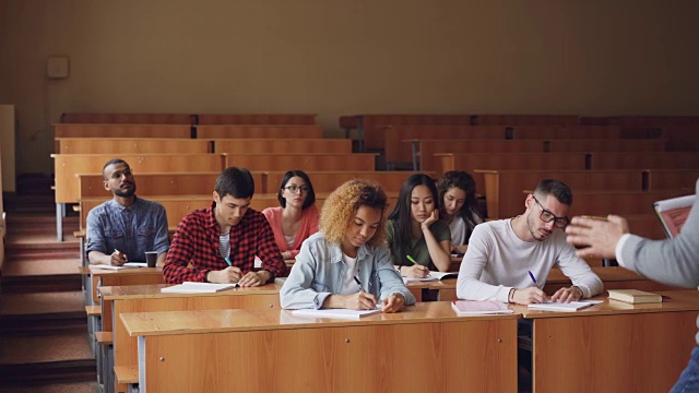 教授正在为一群站在教室里拿着论文的多种族学生朗读演讲，而年轻人则在听他讲课和写作。视频素材