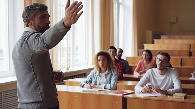 在宽敞的大学教室里，大胡子教授正在读书、说话、打手势，而学生们则坐在桌子旁听、写。视频素材