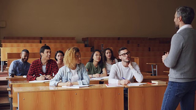 在宽敞的大学讲堂里，导师正在对坐在课桌前的学生讲话，穿着便服的年轻人正在提问。教育与大学理念。视频素材