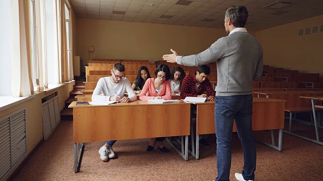 勤奋的学生坐在课桌前写试卷，而戴眼镜的年轻人正在问问题与老师交谈。教育、知识和人的观念。视频素材