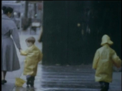 1957年，比利时布鲁塞尔，一名妇女把蹒跚学步的孩子放下，和他一起走，孩子们穿着黄色塑料雨具和一名妇女一起走在人行道上视频素材