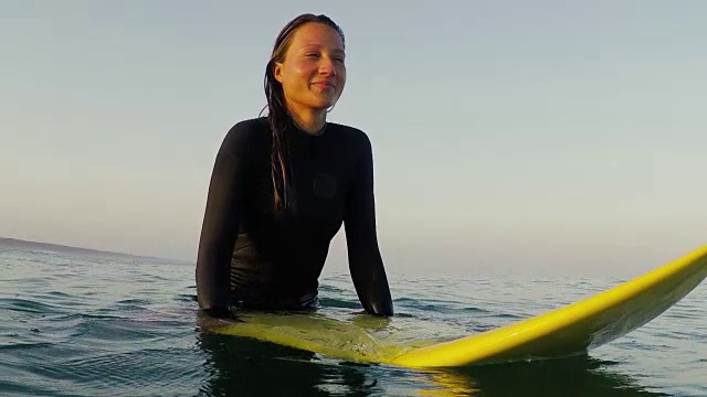 美丽的肖像，微笑的女性冲浪者坐在冲浪板在日出在冲浪休息和海浪在大西洋海岸在法国南部海岸。视频素材