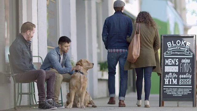 朋友们和金毛猎犬坐在市中心的店面外，人们从人行道上走过。视频素材