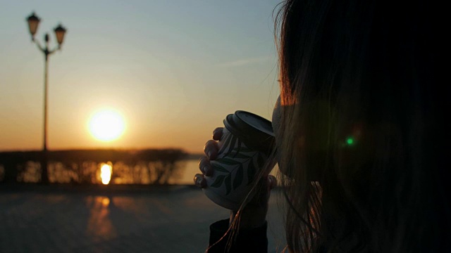 一个女人拿着咖啡杯坐在河堤上，看着清晨从河上升起的太阳视频素材画面1