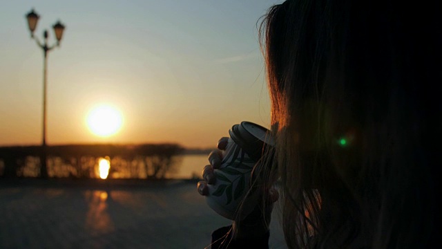 一个女人拿着咖啡杯坐在河堤上，看着清晨从河上升起的太阳视频素材画面4