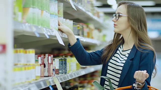 在超级市场:美丽的年轻女子浏览在商店的罐头食品部分。她的购物篮里装满了健康食品。视频素材