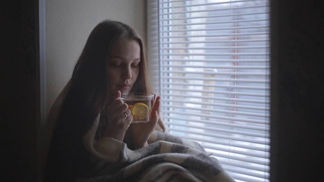 一个生病的漂亮女人感冒了，喝着柠檬热茶，坐在一个乡村房子的窗户在冬天视频素材画面1