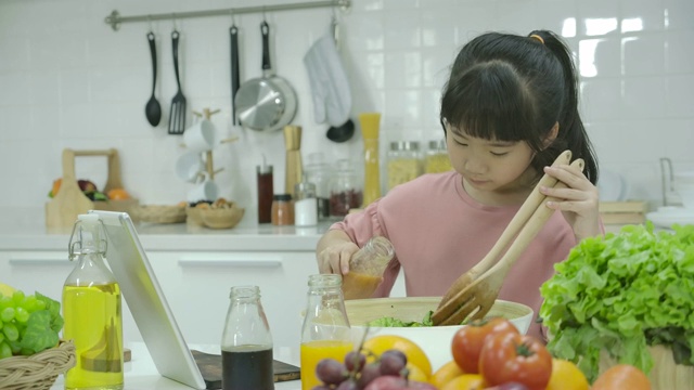 一个亚洲小女孩在家厨房做饭的时候在平板电脑上看沙拉食谱。视频素材