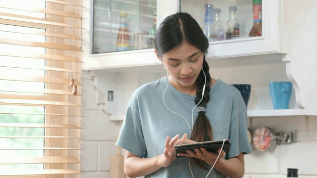 年轻美丽的亚洲女人站在厨房里使用平板电脑和耳机，人们和科技，Z一代的生活方式视频素材