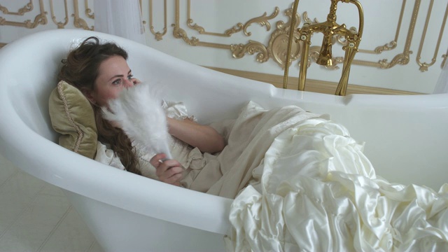 美丽的年轻女子躺在浴缸里，在柔软的枕头上，用手拿着羽毛扇讲电话视频素材画面2