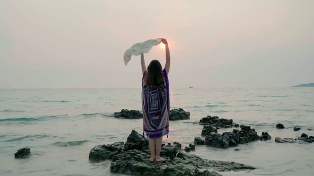 亚洲快乐的女人走在海滩上，在日落时拿着随风飘扬的披肩视频素材