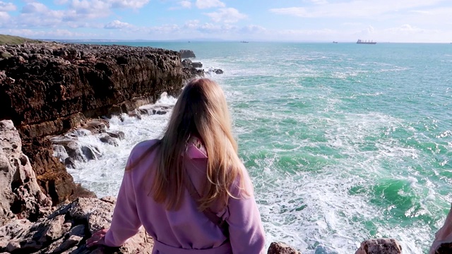 在晴朗的天气里，强烈的海浪撞击着岩石。纯粹的力量，自然力量的展示。强大的能量。海洋力量对抗地球。年轻的金发女人坐在海边的悬崖上看着海浪视频素材