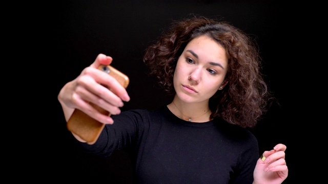 年轻迷人的白人女性的特写肖像自拍手机上的背景孤立在黑色视频素材