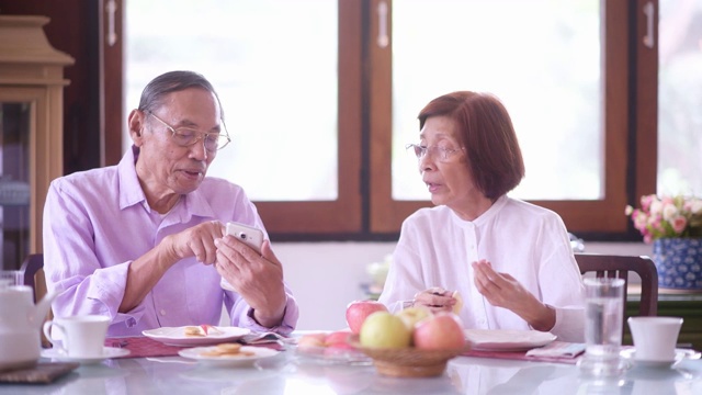 亚洲老年夫妇在家里吃水果时使用智能手机。快乐的老年人生活理念视频素材