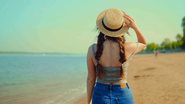 年轻女子沿着海边散步，享受着。一位戴着帽子的女士沿着海滩走，用手碰了碰帽子。后视图。视频素材