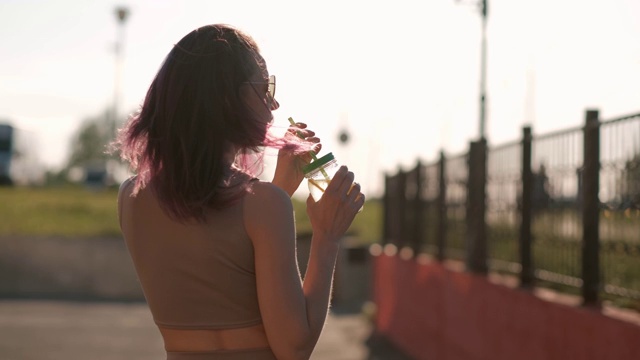 年轻漂亮的女人戴着太阳镜，在城市的街道上喝着用吸管从玻璃罐里舀出的鸡尾酒。视频素材