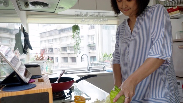 一个女人正在厨房里准备晚餐，看着平板上的食谱视频素材