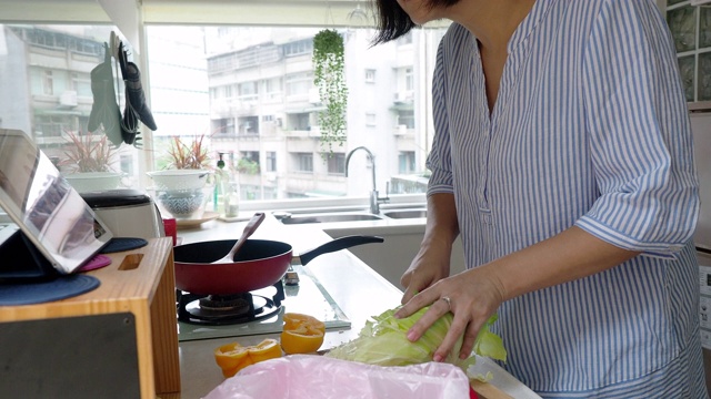 一个女人正在厨房里准备晚餐，看着平板上的食谱视频素材