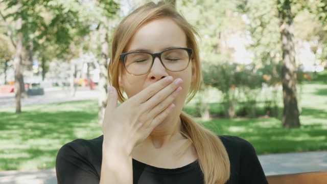 戴眼镜的漂亮女人看着镜头打了个哈欠。迷人的女孩坐在城市公园的长椅上打呵欠视频素材