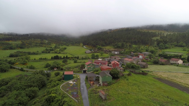 航拍:在多雾的天气里，无人机接近村庄里的房屋和田野，山上的绿色景观映衬着天空——西班牙加利西亚视频素材