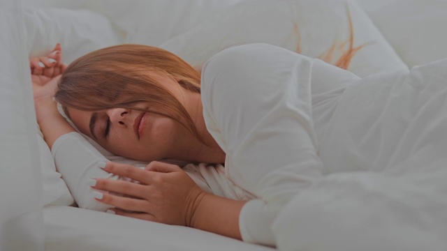 漂亮的女孩在床上盖着白毯子睡得很香。舒适的房间，床上有白色枕头。肖像的观点。视频素材