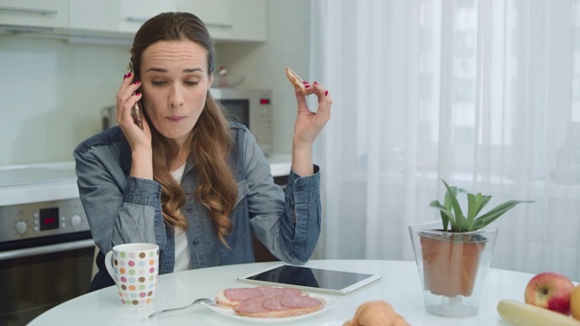 在电话里微笑的女人。家庭主妇在家吃三明治和咖啡。视频素材