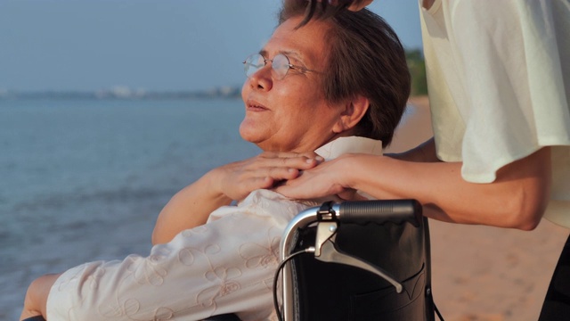 微笑快乐的亚洲资深母亲与她的成年女儿拥抱在海滩上的肖像。快乐的女儿在一起的时候牵着看护者的手。老年假期，假期，关怀，退休，家庭观念，不同能力的旅行视频素材画面4