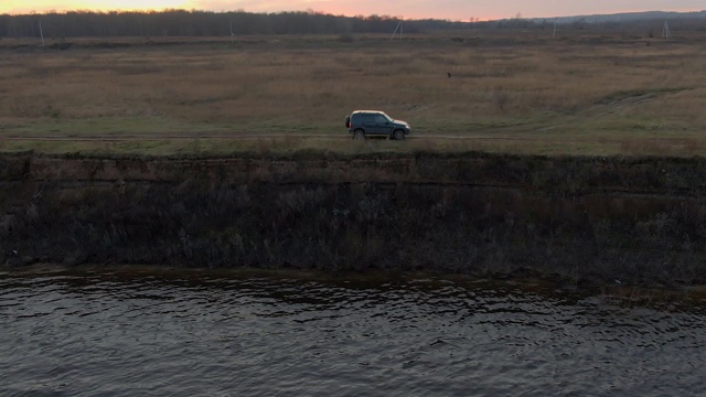 越野道路上的黑色跨界车行驶在河岸上的金色日落景观。在傍晚的阳光下，越野车沿着海边行驶。视频素材