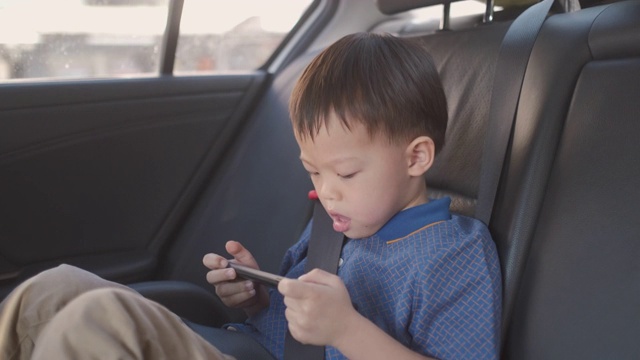 亚洲小男孩坐在婴儿座椅上看来自智能手机的视频，孩子们在车里用智能手机视频素材