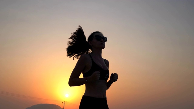 活泼苗条的女孩慢跑在乡村道路与傍晚的天空在背景。女运动员在做跑步训练。年轻的运动妇女在户外工作日落时间。健康积极的生活方式。低的观点视频素材