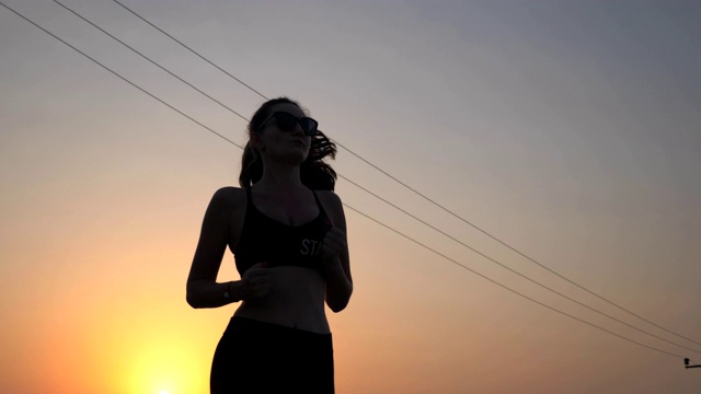 活泼苗条的女孩慢跑在乡村道路与傍晚的天空在背景。女运动员在做跑步训练。年轻的运动妇女在户外工作日落时间。健康积极的生活方式。低的观点视频素材