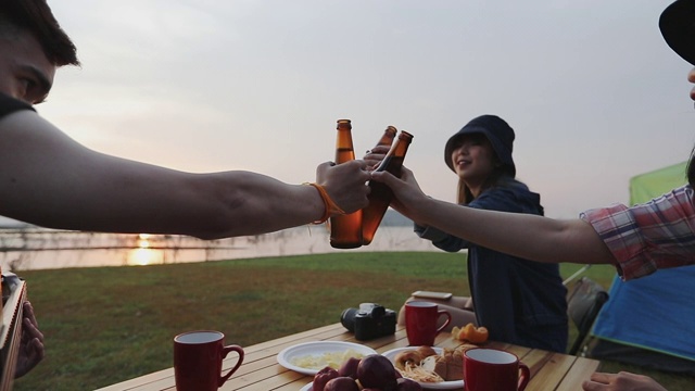 一群亚洲朋友在夏天喝着酒精啤酒，玩着吉他，在日落时分在湖边露营视频素材