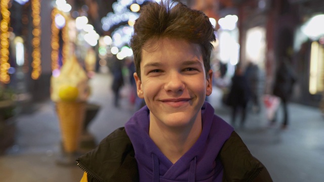 漂亮的少年微笑，看着相机在繁忙的城市街道与城市灯光在晚上。UHD 4 k视频素材画面1