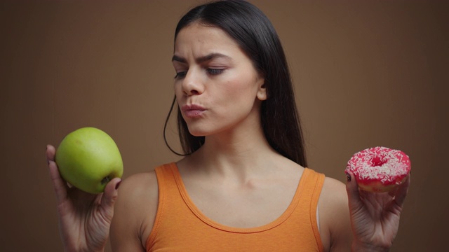 深褐色头发的女人在苹果和甜甜圈之间选择孤立的棕色背景视频素材