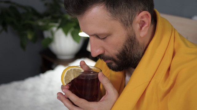 那家伙在喝红茶。有魅力的年轻人病了，坐在床上喝柠檬茶。感冒、流感、喉咙痛、流鼻涕、急性呼吸道疾病等概念。慢动作特写视频素材