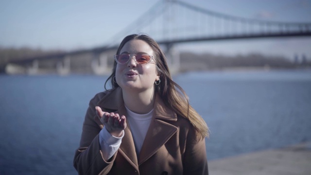 正面的白人胖女孩的肖像送飞吻相机。年轻开朗的女人戴着太阳镜在河边享受阳光明媚的秋日。快乐，生活方式，幸福，休闲。视频素材