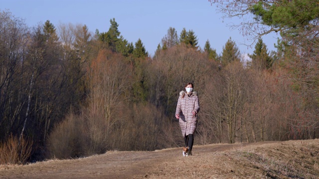 年轻女子穿着保暖外套，戴着口罩，在乡间小路上遛狗。在一些国家，新冠肺炎疫情暴发期间，外出必须佩戴口罩视频素材