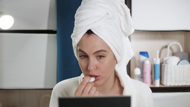 女孩画的嘴唇。有魅力的女人白色毛巾和浴袍坐在浴室对面的镜子和油漆她的嘴唇卫生口红。美容师，在家单独化妆的概念。特写镜头视频素材