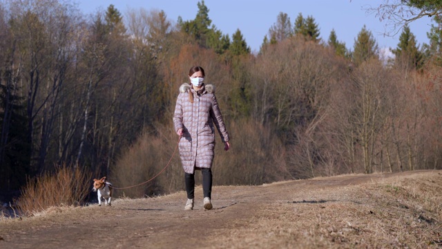 年轻女子穿着保暖外套，戴着口罩，在乡间小路上遛狗。在一些国家，新冠肺炎疫情暴发期间，外出必须佩戴口罩视频素材