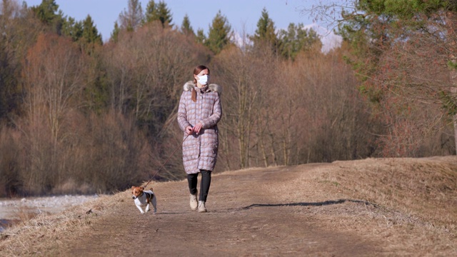 年轻女子穿着保暖外套，戴着口罩，在乡间小路上遛狗。在一些国家，新冠肺炎疫情暴发期间，外出必须佩戴口罩视频素材