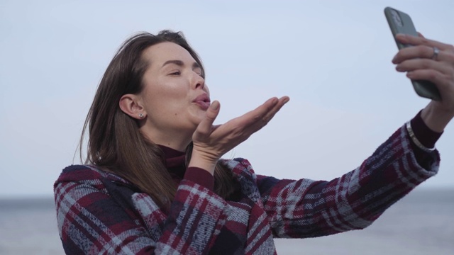 欢快的黑发女孩的特写镜头在自拍相机发送飞吻。年轻的白人女性正在河边拍照。快乐，生活方式，休闲，千禧一代。视频素材