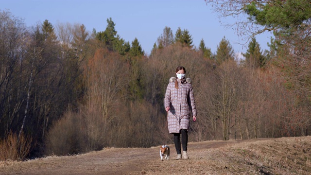 年轻女子穿着保暖外套，戴着口罩，在乡间小路上遛狗。在一些国家，外出时必须佩戴口罩视频素材