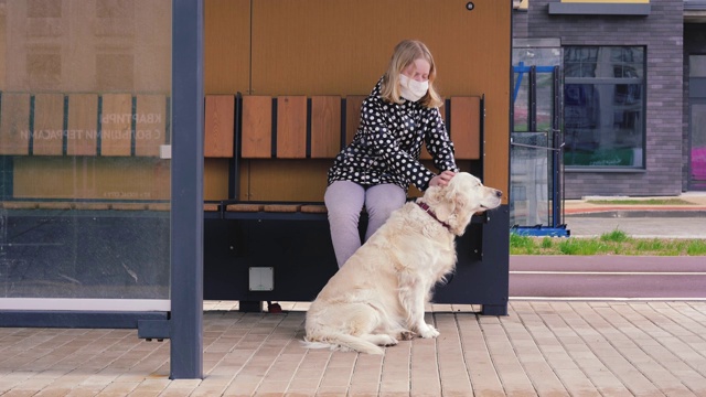 冠状病毒在城市流行。一个戴着防护面具的女孩和一只狗坐在公共交通站视频素材