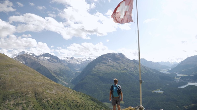 一名男性徒步旅行者站在山顶上俯瞰瑞士令人惊叹的阿尔卑斯风景。瑞士国旗在风中飘扬视频素材